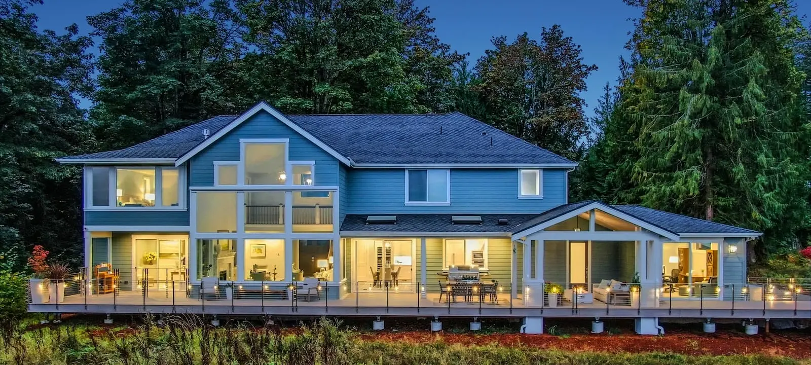 A custom home at dusk with deck and landscape lighting on and exterior shades above the covered porch